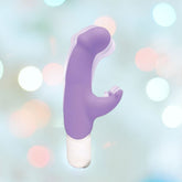 A VeDO Joy Vibe G-Spot Rabbit Vibrator, featuring a purple silicone finish and a curved shaft with an additional external stimulation arm, is set against a blurry background adorned with soft, multicolored bokeh lights. The white base of this VeDO vibrator promises double stimulation for an enhanced experience.